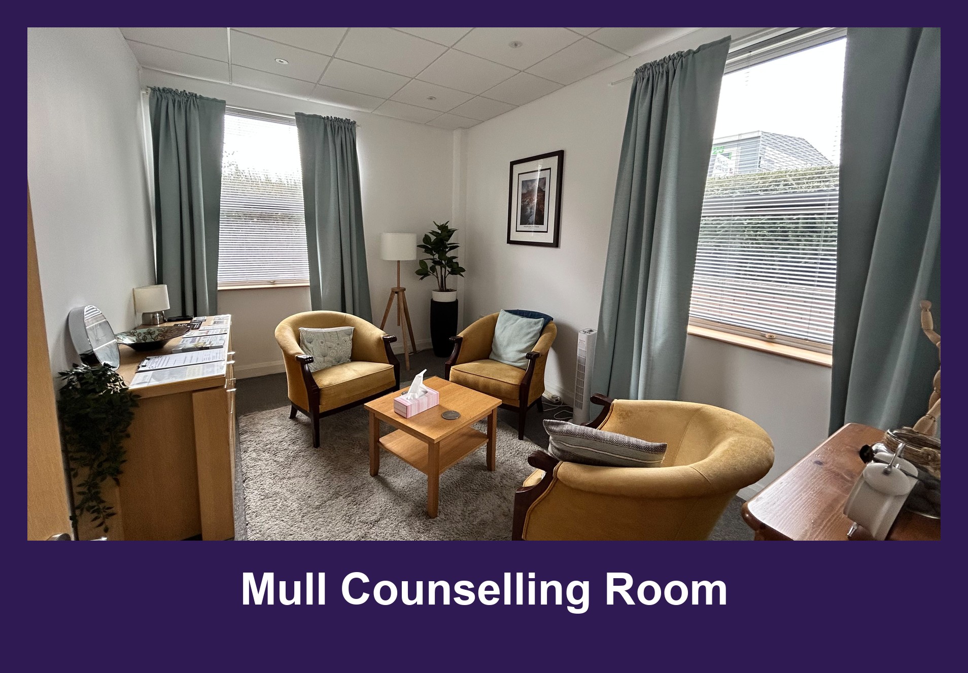 The counselling room at The Bridge Wellbeing Hub's Mull Counselling Room: three mustard-yellow armchairs around a low wooden table on a grey rug, sage-green curtains at two windows, a floor lamp, a framed picture and a potted plant.