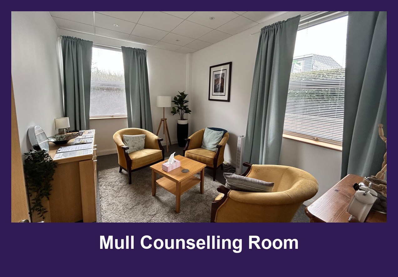 The counselling room at The Bridge Wellbeing Hub's Mull Counselling Room: three mustard-yellow armchairs around a low wooden table on a grey rug, sage-green curtains at two windows, a floor lamp, a framed picture and a potted plant.